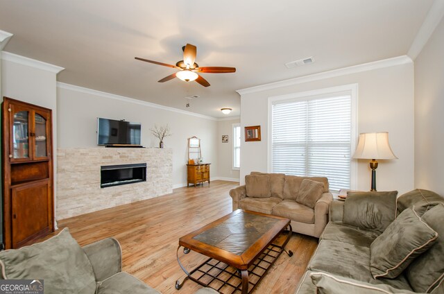 living area featuring a stone fireplace, ornamental molding, a ceiling fan, visible vents, and wood finished floors