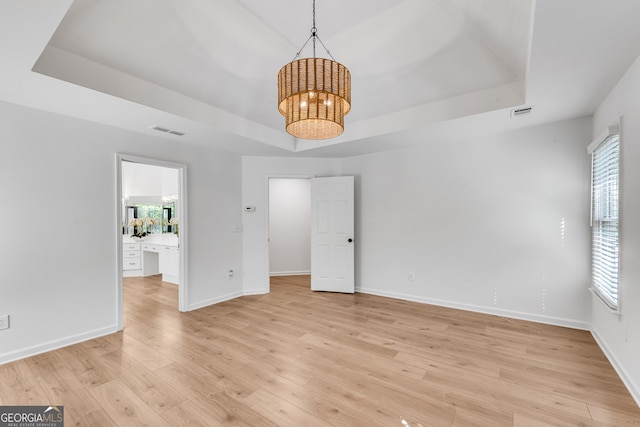 empty room with a raised ceiling, a chandelier, healthy amount of natural light, and light wood-style floors