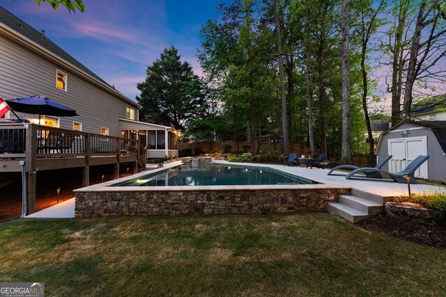pool at dusk featuring a storage unit, a sunroom, a fenced backyard, and a deck
