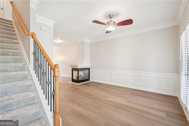 unfurnished living room with a multi sided fireplace, a ceiling fan, crown molding, stairway, and light wood-style flooring