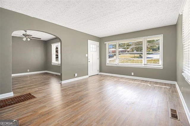unfurnished living room featuring ceiling fan, arched walkways, a textured ceiling, visible vents, and wood finished floors