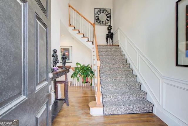 staircase featuring a decorative wall, wainscoting, a towering ceiling, and wood finished floors