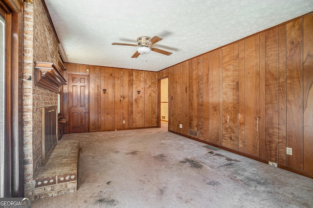 empty room featuring a textured ceiling, ceiling fan, wood walls, a brick fireplace, and visible vents
