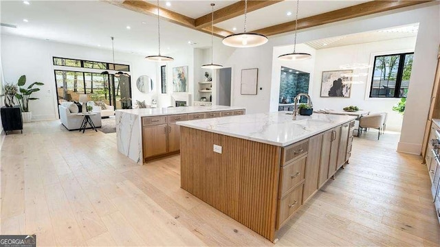 kitchen with a sink, a wealth of natural light, light wood-type flooring, beamed ceiling, and a large island with sink
