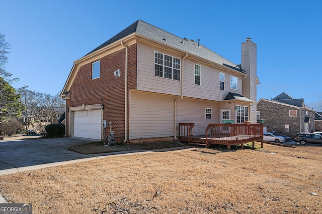 rear view of house with a wooden deck, roof with shingles, an attached garage, a chimney, and concrete driveway