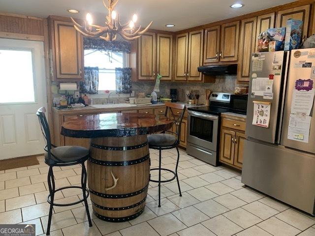 kitchen featuring an inviting chandelier, under cabinet range hood, brown cabinetry, stainless steel appliances, and backsplash