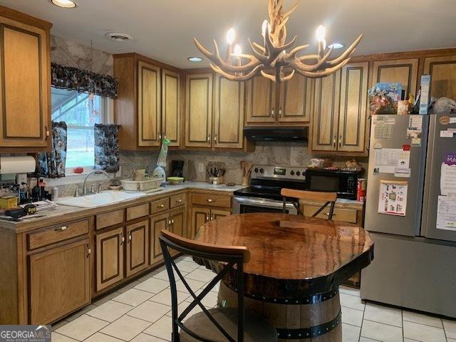 kitchen with a sink, appliances with stainless steel finishes, a chandelier, under cabinet range hood, and light tile patterned flooring