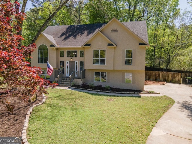 raised ranch featuring fence, a shingled roof, a front lawn, and stucco siding