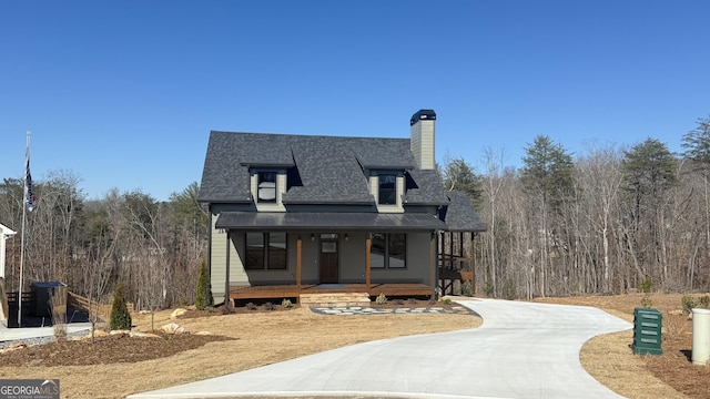 view of front of house with concrete driveway, roof with shingles, a chimney, a wooded view, and a porch