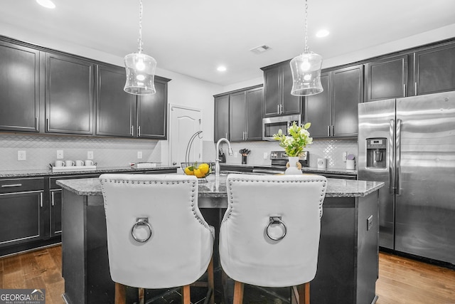 kitchen with tasteful backsplash, decorative light fixtures, appliances with stainless steel finishes, an island with sink, and light stone countertops