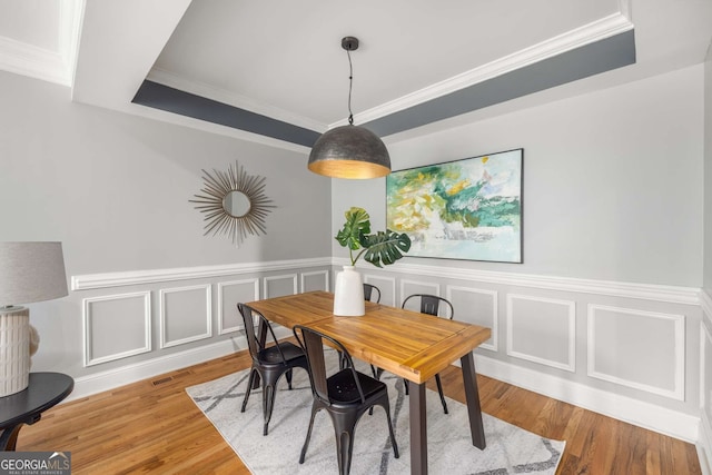 dining space featuring a raised ceiling, a decorative wall, crown molding, wood finished floors, and a wainscoted wall