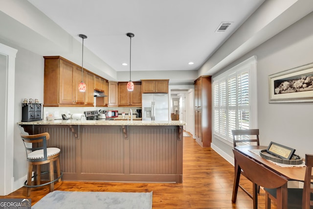 kitchen with light stone counters, light wood finished floors, brown cabinetry, a peninsula, and refrigerator with ice dispenser