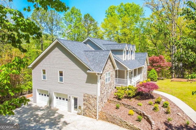 view of side of property featuring concrete driveway, stone siding, an attached garage, a shingled roof, and covered porch