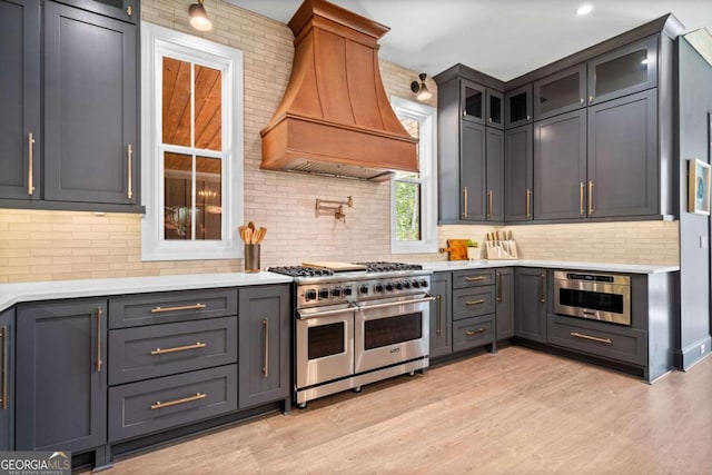 kitchen featuring glass insert cabinets, stainless steel appliances, custom range hood, light countertops, and backsplash