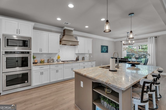 kitchen featuring tasteful backsplash, premium range hood, a center island with sink, stainless steel appliances, and a sink
