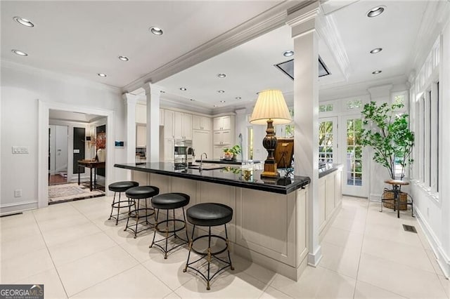kitchen with ornamental molding, light tile patterned flooring, dark countertops, a kitchen bar, and decorative columns