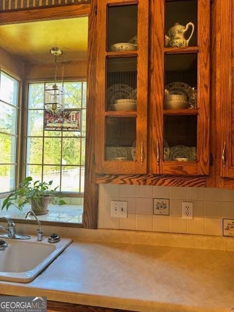 interior details with glass insert cabinets, hanging light fixtures, a sink, brown cabinetry, and backsplash