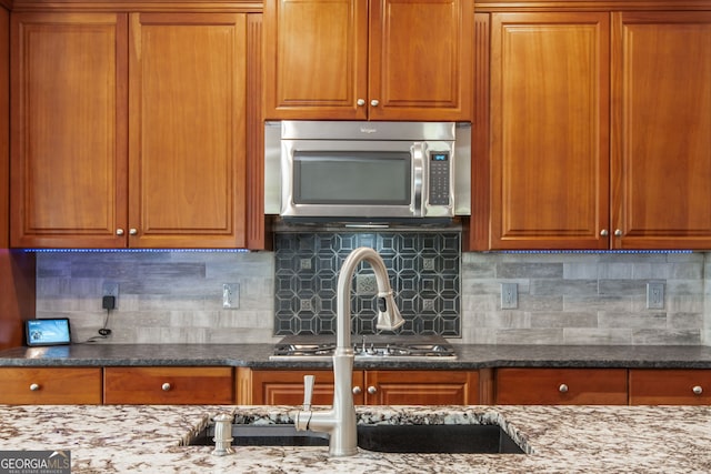 kitchen with brown cabinets, backsplash, stainless steel microwave, and dark stone countertops