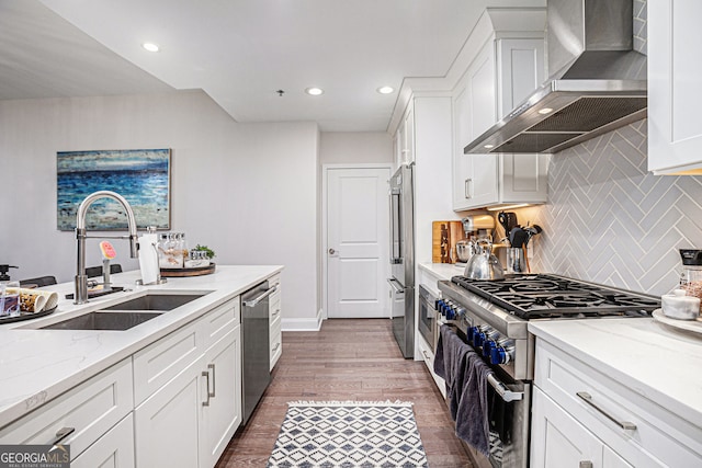 kitchen featuring wall chimney range hood, premium appliances, a sink, backsplash, and white cabinetry