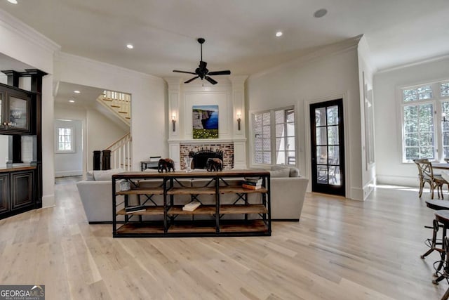 living room featuring ornamental molding, stairway, light wood-style flooring, a ceiling fan, and a brick fireplace