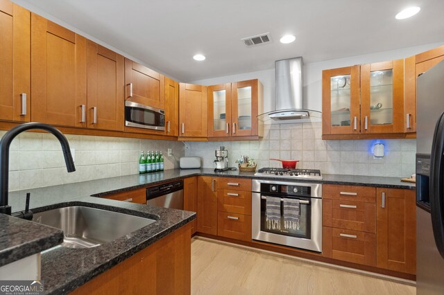 kitchen with dark stone countertops, a sink, wall chimney range hood, brown cabinetry, and stainless steel appliances