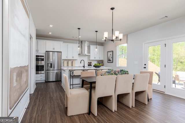 kitchen featuring wall chimney range hood, a center island with sink, appliances with stainless steel finishes, dark wood-type flooring, and visible vents