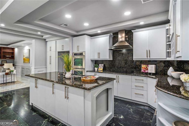 kitchen featuring stainless steel double oven, a tray ceiling, white cabinetry, and wall chimney range hood