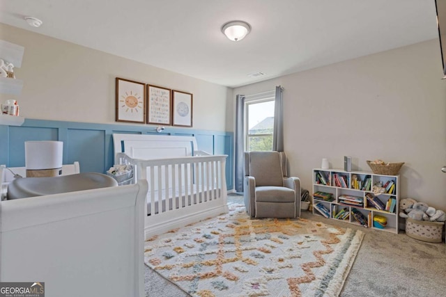 carpeted bedroom with a decorative wall, visible vents, a crib, and wainscoting