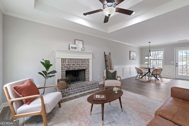 living area with a brick fireplace, wood finished floors, ceiling fan, crown molding, and a raised ceiling