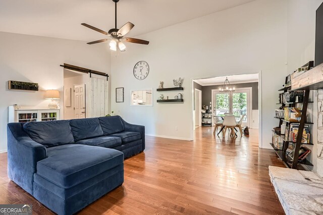living room featuring wood finished floors, baseboards, high vaulted ceiling, a barn door, and ceiling fan with notable chandelier