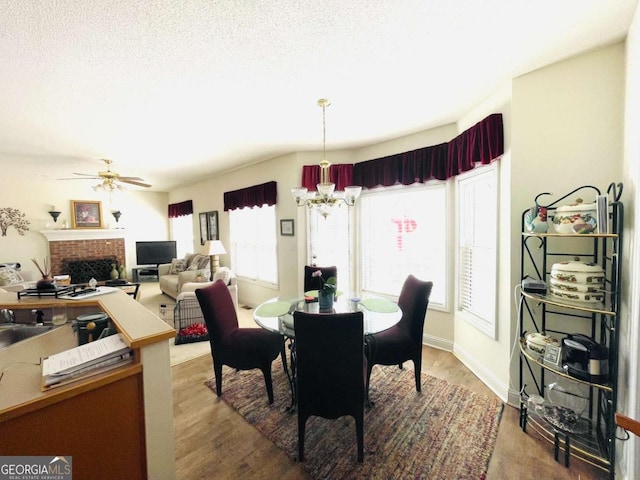 dining space featuring a textured ceiling, wood finished floors, a brick fireplace, and ceiling fan with notable chandelier