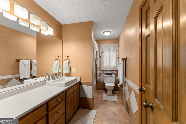 full bath featuring a wainscoted wall, toilet, vanity, tile walls, and a textured ceiling