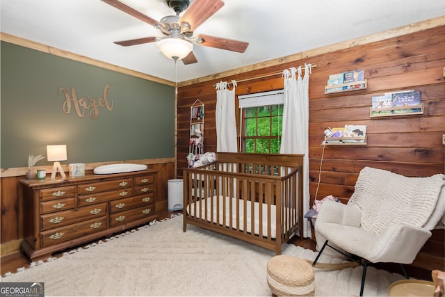 bedroom featuring a crib, wooden walls, wood finished floors, a ceiling fan, and wainscoting