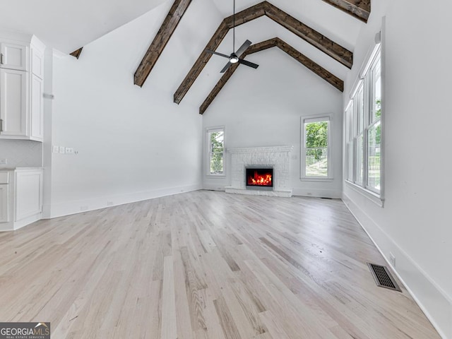 unfurnished living room with high vaulted ceiling, ceiling fan, light wood-style flooring, beam ceiling, and a stone fireplace