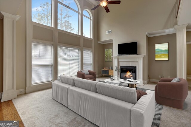living area featuring a glass covered fireplace, ceiling fan, a towering ceiling, visible vents, and decorative columns