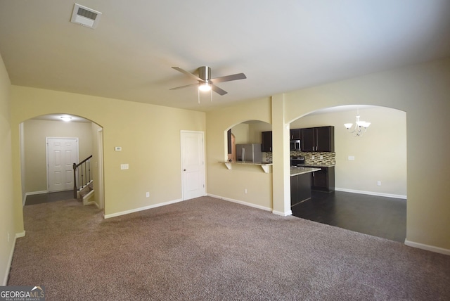 unfurnished living room featuring dark colored carpet, baseboards, visible vents, ceiling fan with notable chandelier, and stairway