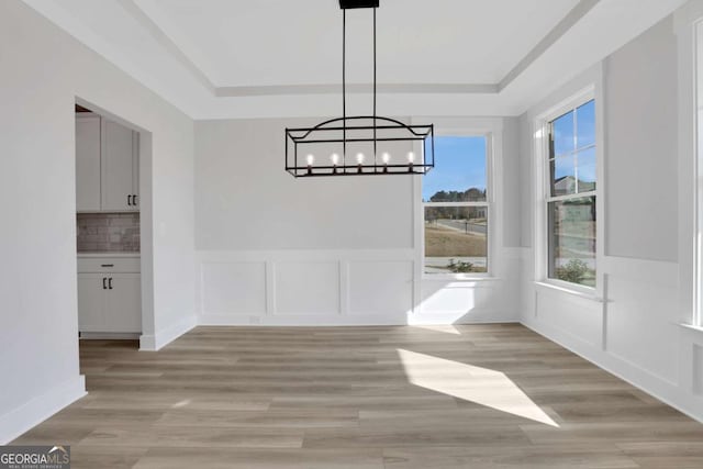 unfurnished dining area with a decorative wall, a notable chandelier, a tray ceiling, and light wood-style flooring