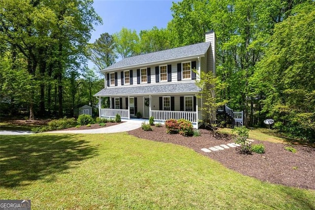 colonial inspired home with a chimney, covered porch, a shingled roof, a shed, and a front yard