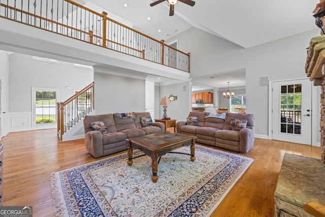 living room with ceiling fan with notable chandelier, light wood-style flooring, high vaulted ceiling, and stairway