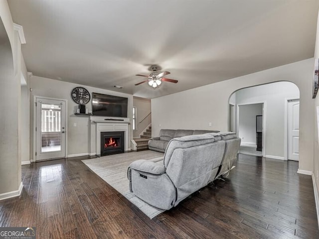living room featuring arched walkways, dark wood-style flooring, baseboards, ceiling fan, and a lit fireplace
