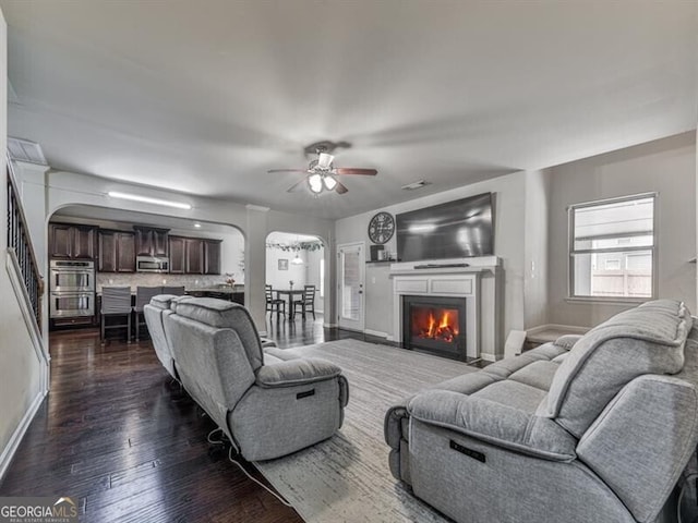 living room with a ceiling fan, arched walkways, dark wood-type flooring, baseboards, and a glass covered fireplace