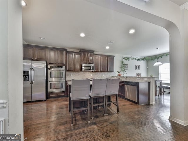 kitchen featuring a peninsula, arched walkways, dark brown cabinets, decorative backsplash, and appliances with stainless steel finishes