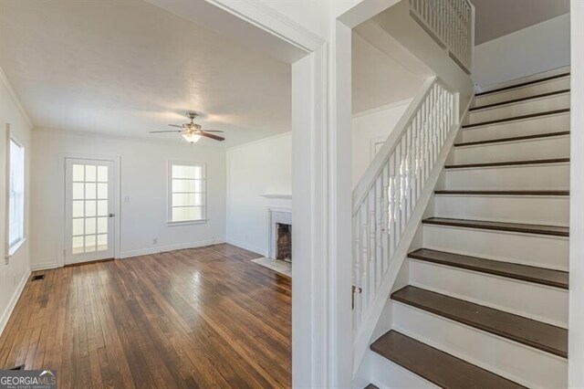 stairway featuring hardwood / wood-style flooring, ceiling fan, baseboards, a fireplace with flush hearth, and crown molding