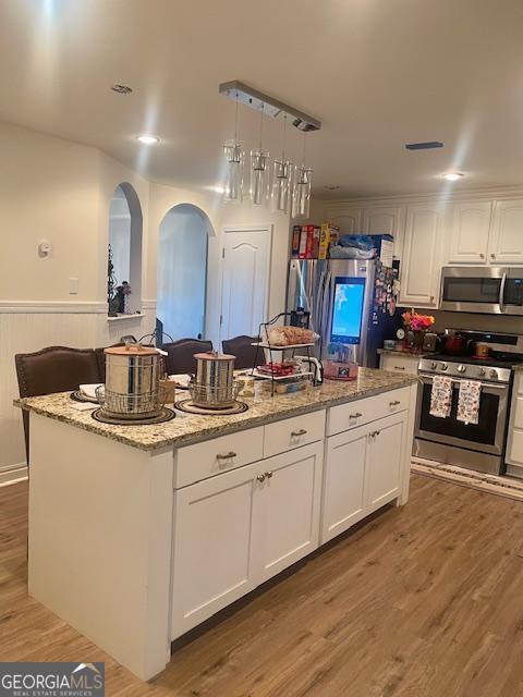 kitchen with stainless steel appliances, arched walkways, a center island, white cabinetry, and a wainscoted wall