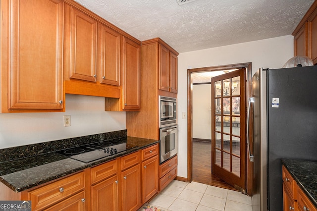 kitchen with appliances with stainless steel finishes, light tile patterned floors, brown cabinets, dark stone counters, and a textured ceiling