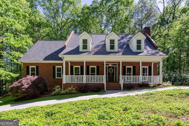 cape cod house with a chimney, a porch, a front yard, brick siding, and roof with shingles
