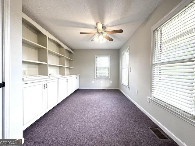 unfurnished office featuring visible vents, dark carpet, baseboards, a textured ceiling, and a ceiling fan