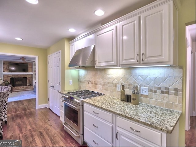 kitchen with wall chimney range hood, white cabinetry, a brick fireplace, and stainless steel stove