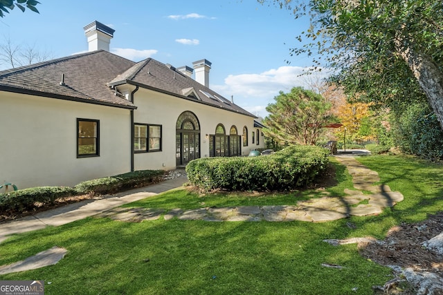 rear view of house featuring a lawn, a chimney, stucco siding, and roof with shingles