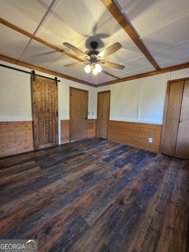 unfurnished bedroom with wooden walls, a barn door, a ceiling fan, and dark wood-style flooring